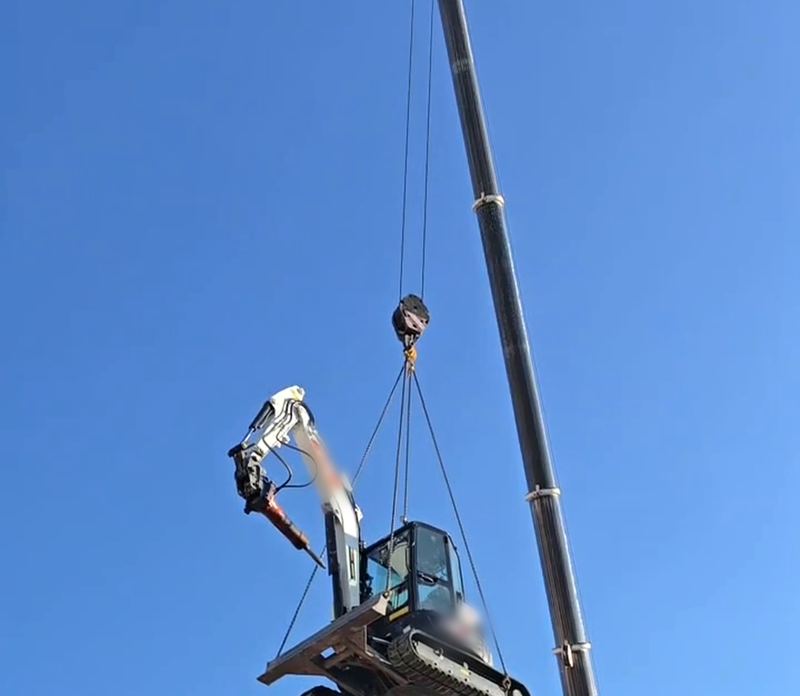Grúa elevando una miniexcavadora por encima del tejado de una vivienda para acceder a la parcela