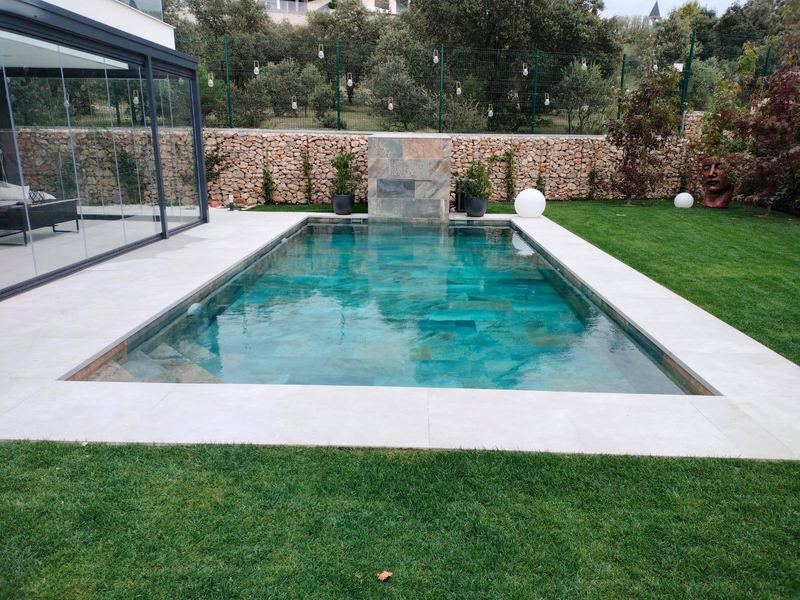 Piscina de obra en jardín con revestimiento porcelánico efecto piedra verde, cascada revestida de piedra al fondo, coronación clara y césped alrededor