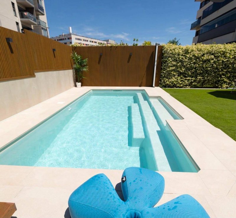 Piscina de obra con escalera de obra ancha integrada en un lateral, coronación porcelánica clara, césped artificial y cerramiento de lamas de madera